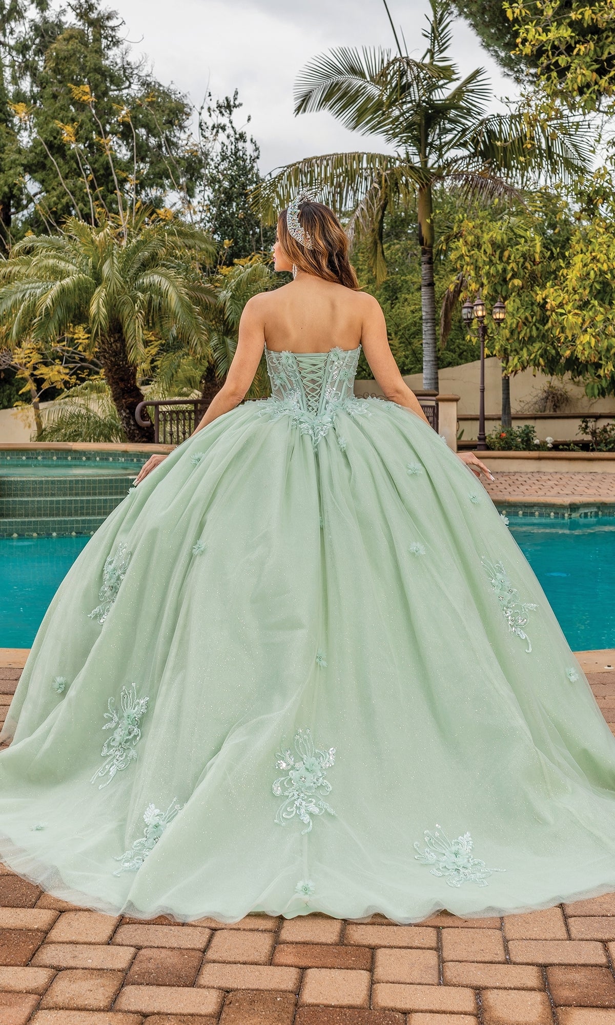 A woman stands by a pool, facing away from the camera, in the Quinceanera Dress 1893 By Dancing Queen. The sage green gown features a strapless sweetheart neckline, full skirt, and floral embroidery with palm trees and greenery behind her.